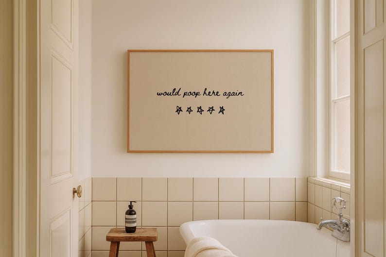 May include: A framed print with the text "would poop here again" and star graphics hangs above a bathtub. The bathroom has a neutral color palette with a wooden stool and a soap dispenser.