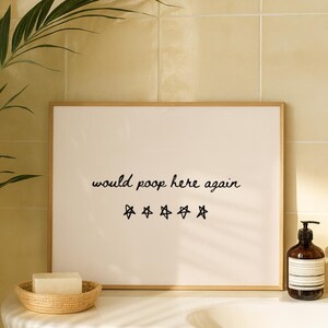 May include: A framed print with the text "would poop here again" and five star symbols. A brown soap dispenser, a small hourglass, and a woven basket with a bar of soap are also visible.