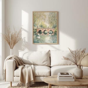 May include: A framed watercolor print of a bridge over a river with two swans. The bridge is made of red brick and has three arches. The print is in a light wood frame and hangs above a white sofa.