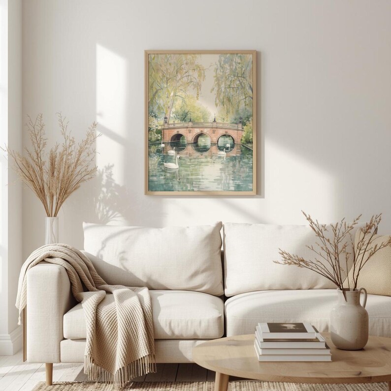 May include: A framed watercolor print of a bridge over a river with two swans. The bridge is made of red brick and has three arches. The print is in a light wood frame and hangs above a white sofa.