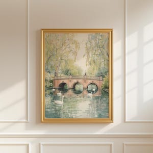 Puede incluir: Una pintura de acuarela enmarcada representa un sereno paisaje acuático con un puente de piedra y cisnes. El puente tiene tres arcos y se refleja en el agua. La pintura está en un marco dorado y cuelga de una pared blanca.
