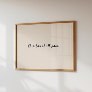 May include: A framed print with the phrase "this too shall pass" in black script on a cream-colored background. The frame is made of light-colored wood, and the print is displayed on a white wall.