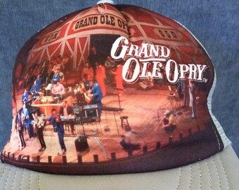 May include: A brown and white trucker hat with a photo of a stage with musicians performing. The text "Grand Ole Opry" is printed in white on the hat.
