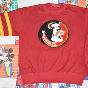 Può includere: Felpa rossa con un grande logo ricamato Seminole State sul davanti. Il logo è una testa di nativo americano con un design rosso e giallo. La felpa ha strisce gialle e rosse sulle maniche.