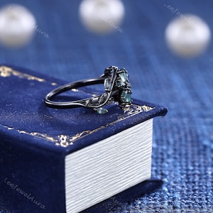 May include: A black ring featuring a floral design and green gemstones. The ring rests on a blue book with gold edging. The ring's band has a detailed leaf pattern.