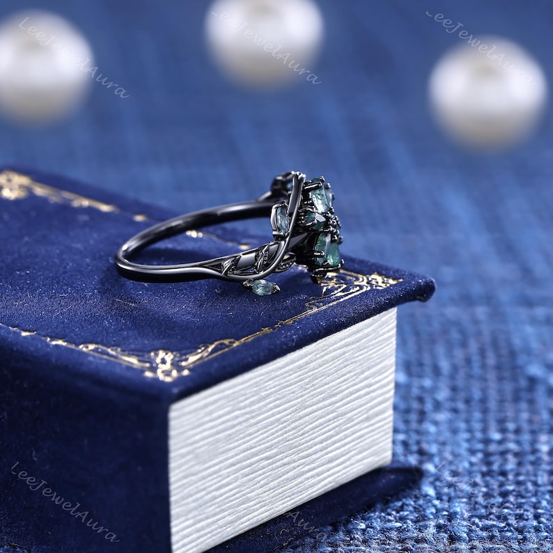 May include: A black ring featuring a floral design and green gemstones. The ring rests on a blue book with gold edging. The ring's band has a detailed leaf pattern.