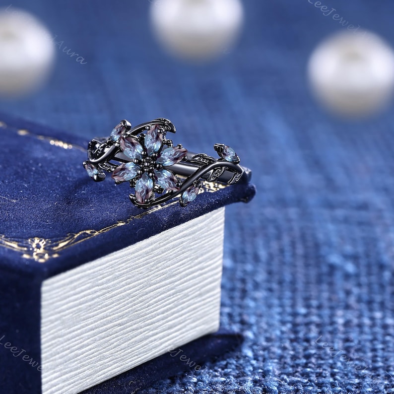 May include: A black floral ring with a central flower design and smaller leaf accents. The ring features a cluster of blue and purple gemstones, set against a dark metal band. The ring is displayed on a blue surface.