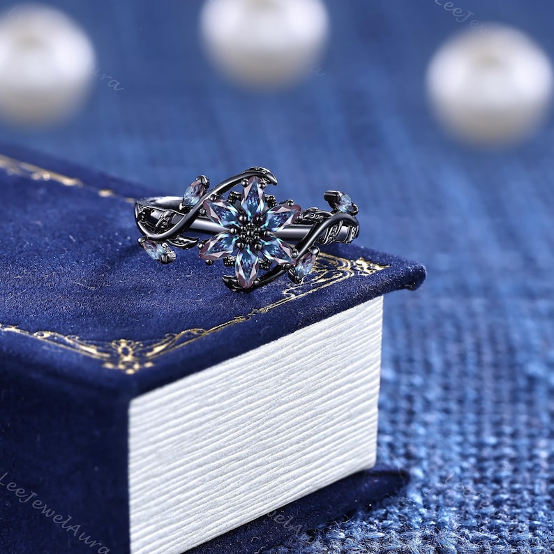 May include: A black metal ring with a floral design and a central blue-green gemstone. The ring has a delicate, vine-like band with small blue gemstones. The ring is set against a dark blue background.