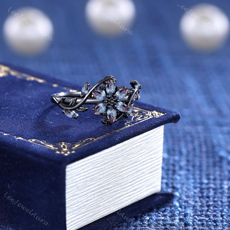 May include: A black floral ring with blue and purple gemstones. The ring has a flower design with detailed leaves and vines. The ring is set on a blue book.