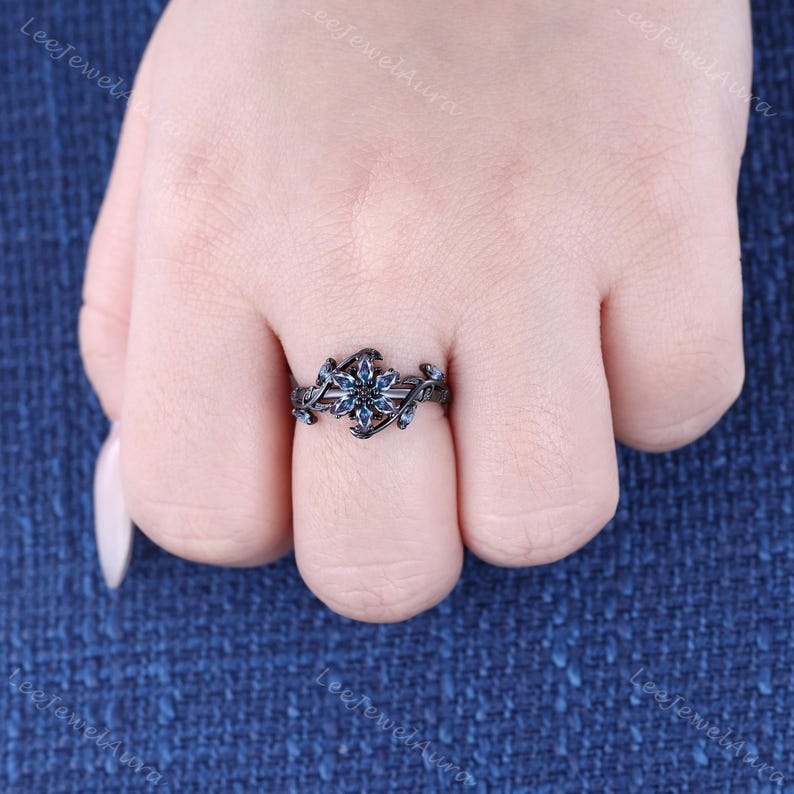 May include: A black floral ring with blue gemstones. The ring features a central flower design with surrounding leaf and vine details. The gemstones are a deep blue colour, set against the black metal.