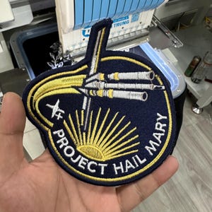 Op de afbeelding: Een ronde geborduurde patch met een donkerblauwe achtergrond, een geel zonnestraalontwerp aan de onderkant en de tekst PROJECT HAIL MARY in het wit. Een zilveren en gele ruimteschipafbeelding strekt zich uit vanaf het midden naar boven.