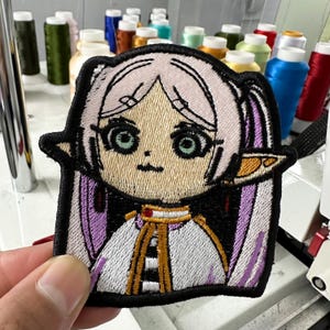 Pode incluir: Um emblema bordado apresentando um personagem estilo anime com cabelo claro, orelhas pontudas e olhos verdes grandes. O personagem veste uma roupa branca e dourada com detalhes em roxo, tudo contornado por uma borda preta espessa.
