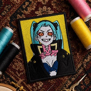 May include: Embroidered patch featuring an anime-style character with blue hair, red eyes, and a sinister grin. The patch has a yellow background and a black border. Sewing supplies, including thread spools and scissors, surround the patch.