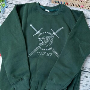 May include: Dark green sweatshirt with white graphic text and illustrations. The design includes two crossed swords, a geometric bear, and the phrases "Silver for Monsters" and "Steel for Humans".