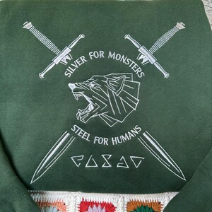 May include: Dark green sweatshirt with white embroidered design. The design includes crossed swords, a wolf head, and the text "SILVER FOR MONSTERS" and "STEEL FOR HUMANS". The bottom of the sweatshirt has a colorful, crocheted pattern.