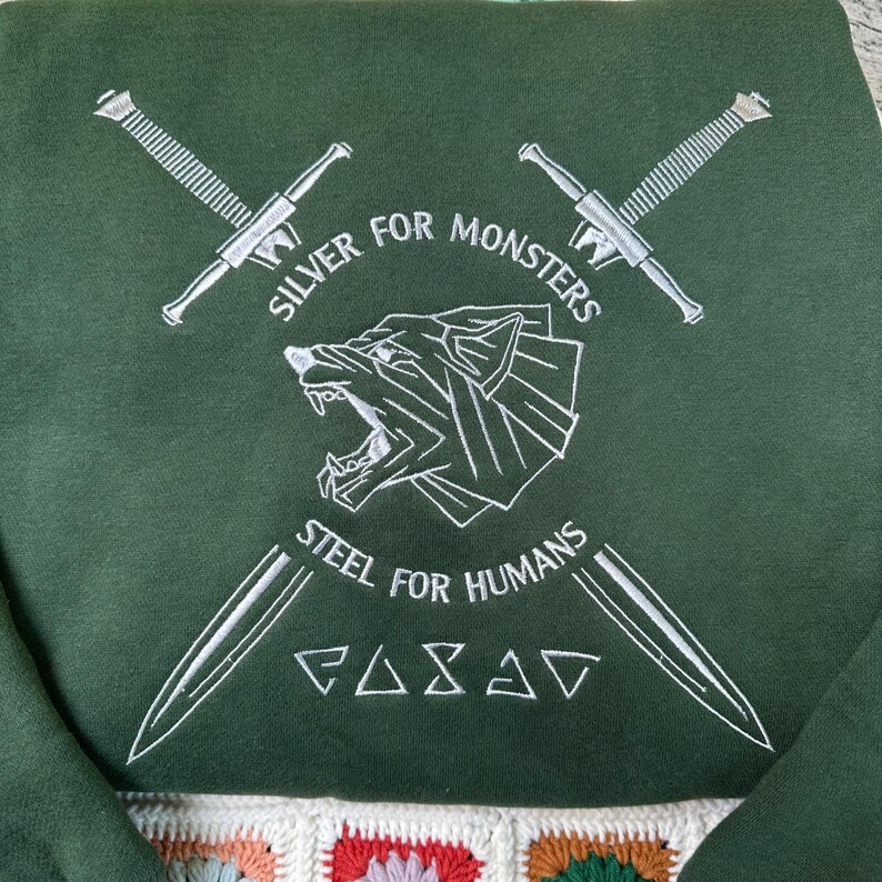 May include: Dark green sweatshirt with white embroidered design. The design includes crossed swords, a wolf head, and the text "SILVER FOR MONSTERS" and "STEEL FOR HUMANS". The bottom of the sweatshirt has a colorful, crocheted pattern.