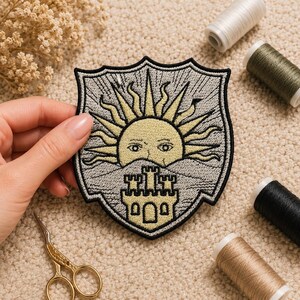 May include: Embroidered patch featuring a shield design with a yellow sun and a castle. The patch has a black border and is on a beige background. Sewing supplies, including thread spools and scissors, are visible.
