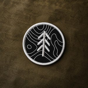 May include: Embroidered patch featuring a white tree design on a black circular background. The tree is surrounded by white contour lines, with a white border. The patch is set against a dark olive green fabric.