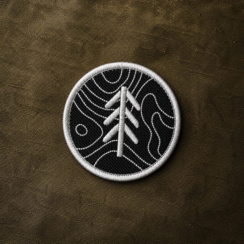 May include: Embroidered patch featuring a white tree design on a black circular background. The tree is surrounded by white contour lines, with a white border. The patch is set against a dark olive green fabric.