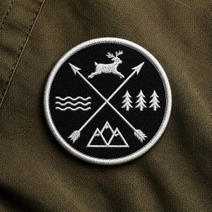 Op de afbeelding: Een ronde, zwarte patch met een wit geborduurd ontwerp. Het ontwerp bevat een hert dat over gekruiste pijlen springt, bergen, bomen en water, allemaal binnen een witte rand. De patch is geplaatst tegen een groene stoffen achtergrond.