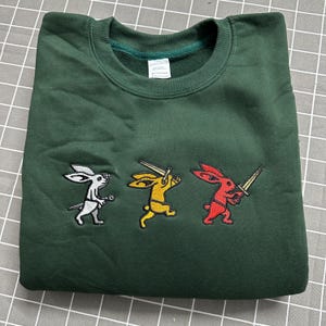 May include: A forest green crewneck sweatshirt featuring an embroidered design of three rabbits in a row. The first rabbit is white, the second is yellow holding a sword, and the third is red holding a sword. The garment has a ribbed collar.