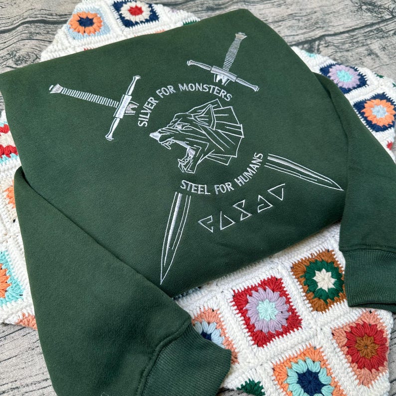 May include: A dark green sweatshirt with white embroidered text and graphics. The design includes crossed swords, a wolf-like creature, and the phrases "SILVER FOR MONSTERS" and "STEEL FOR HUMANS". The sweatshirt is folded on a colorful crocheted blanket.