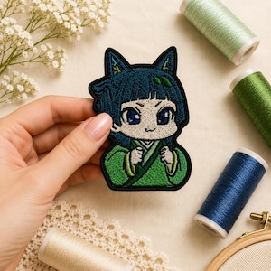 Può includere: Toppa ricamata raffigurante un personaggio in stile anime con capelli blu e orecchie da gatto, in un completo verde. La toppa è tenuta in mano, con rocchetti di filo sullo sfondo. La toppa è alta circa 7.6 cm.