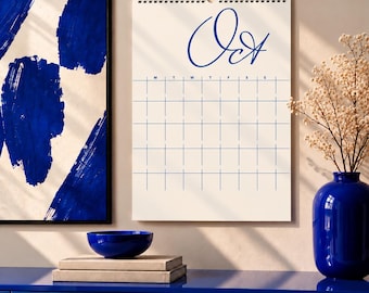 Puede incluir: Un calendario de pared con fondo blanco y líneas de cuadrícula azules, que presenta la palabra Oct en letra cursiva azul en la parte superior. El calendario tiene una espiral en la parte superior y se muestra junto a un cuenco y un jarrón azules.
