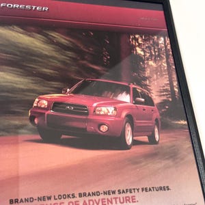 以下が含まれることがあります： ワインレッドのSubaru Forester SUVの広告。車両は、森を背景にした道路に表示されています。テキストには「BRAND-NEW LOOKS. BRAND-NEW SAFETY FEATURES. SENSE OF ADVENTURE.」と書かれています。