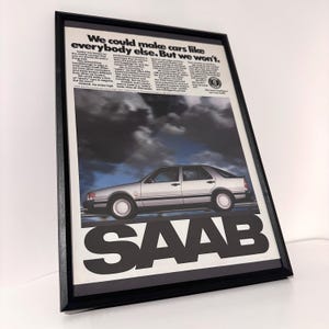 Puede incluir: Anuncio vintage enmarcado de un Saab plateado. La imagen muestra un coche Saab en una carretera con un cielo nublado. El texto dice: "Podríamos hacer coches como todos los demás. Pero no lo haremos." El logotipo de Saab es prominente.