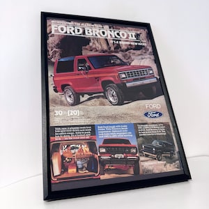 Peut inclure: Publicité encadrée vintage pour un Ford Bronco II. Le SUV rouge est présenté sur un chemin de terre avec le texte "FORD BRONCO II" et "IT'S A BRAND NEW KICK!". Le cadre est noir.
