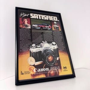 May include: Framed vintage Canon AE-1 camera advertisement. The black frame surrounds a print featuring a Canon AE-1 camera, a race car, and a portrait of a man. The text reads "SATISFIED."