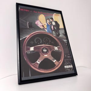 May include: A framed vintage advertisement poster featuring a wood-rimmed MOMO steering wheel in the foreground. The background shows a couple on stairs with the text MOMO - An Independent Style at the top and the MOMO logo in the bottom corner.