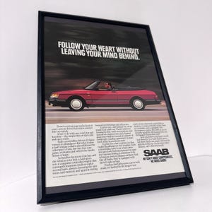 May include: Framed advertisement for a red Saab convertible with the top down, driving on a road. The ad includes the text "FOLLOW YOUR HEART WITHOUT LEAVING YOUR MIND BEHIND." and the Saab logo.