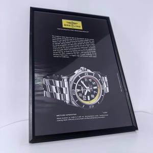 Puede incluir: Anuncio enmarcado de un reloj Breitling Superocean. El reloj tiene una esfera negra con detalles blancos y amarillos, una correa de acero inoxidable y un bisel giratorio unidireccional. El texto publicitario destaca las características del reloj.