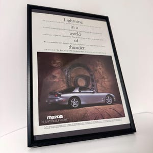 May include: Framed advertisement for a silver Mazda RX-7 sports car. The ad features the car on a brown surface, with text that reads "Lightning in a world of thunder." The Mazda logo and the tagline "It Just Feels Right" are also visible.