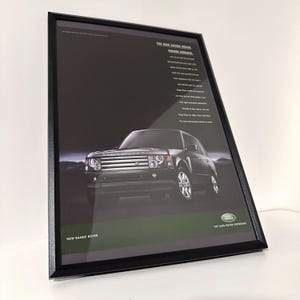 Könnte beinhalten: Gerahmte Werbung für den neuen Range Rover. Das Bild zeigt einen dunkelgrauen SUV mit dem Text "THE NEW RANGE ROVER HIGHER GROUND" und anderem beschreibenden Text. Das Land Rover-Logo ist sichtbar.