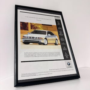 Könnte beinhalten: Gerahmte Werbung mit einem weißen BMW-Limousinen. Die Anzeige enthält Text über zertifizierte Gebrauchtfahrzeuge und das BMW-Logo. Der Rahmen ist schwarz und das Bild ist leicht angewinkelt.