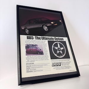 May include: Framed advertisement for BBS wheels. The image features a black car and a close-up of a silver wheel. Text reads "BBS: The Ultimate Option" with a list of car models.