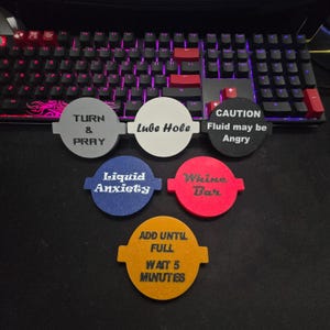 Può includere: Un set di sottobicchieri circolari colorati con frasi umoristiche. I sottobicchieri sono di vari colori, tra cui grigio, bianco, blu, rosso e giallo. Ogni sottobicchiere ha una frase unica come "Lube Hole" e "Liquid Anxiety".
