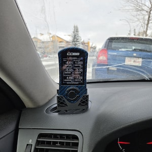 May include: A black and blue digital gauge with the brand name COBB displayed on the screen. The gauge is mounted on a black dashboard inside a car. The screen displays various numerical data.