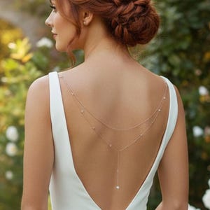Könnte beinhalten: Eine Frau mit rotbraunem Haar, das zu einem Dutt hochgesteckt ist, trägt ein weißes Hochzeitskleid mit tiefem Rückenausschnitt. Eine zarte goldene Körperkette mit kleinen Perlen verziert den Rücken. Der Hintergrund zeigt einen Garten mit weißen Rosen und einem Steinbogen.