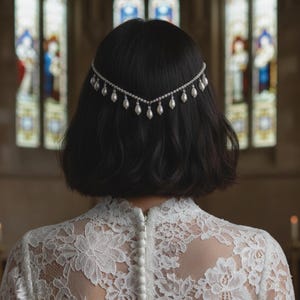 May include: A bride in a white lace wedding dress with long sleeves and a high neckline, featuring a pearl-buttoned back. A delicate headpiece with pearls is worn in the hair. The scene is set in a church.