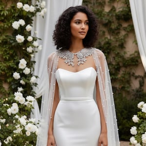 May include: A strapless white wedding dress with a fitted bodice and a flowing skirt. A sheer cape adorned with pearls and a jeweled collar drapes over the shoulders. The background features white roses and greenery.