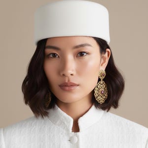 May include: A woman wearing a white hat and a white jacket. She is also wearing gold-colored filigree earrings with a central red stone. The earrings are ornate and dangle from her ears. The background is a neutral beige color.