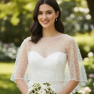May include: A bride wearing a white wedding dress with a sheer, dotted capelet. The capelet is adorned with small, evenly spaced pearls. She is holding a bouquet of white flowers, smiling in front of a blurred green background.