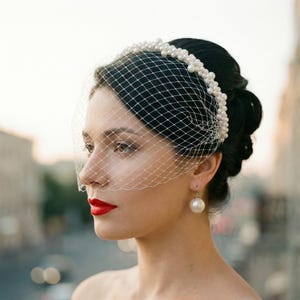 Op de afbeelding: Een vrouw draagt een witte vogelkooissluier en een parelhoofdband. De sluier is bevestigd aan een hoofdband versierd met meerdere parels. De vrouw draagt ook pareloorbellen en rode lippenstift. De achtergrond is wazig.