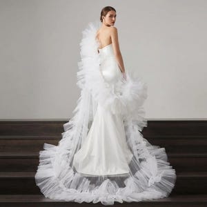 Op de afbeelding: Een witte, strapless trouwjurk met een zeemeermin-silhouet. De jurk heeft een opvallende, gefranjerde tule overlay die uitloopt in een lange sleep. De bruid staat op een donkere houten trap.