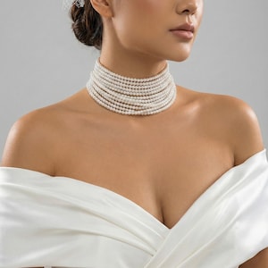 Könnte beinhalten: Ein mehrreihiges Perlen-Choker-Halsband. Die Halskette besteht aus zahlreichen Strängen kleiner, runder, weißer Perlen. Das Model trägt ein weißes schulterfreies Kleid.