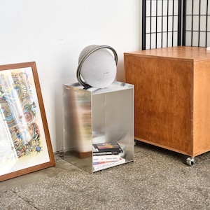 May include: A modern, silver-colored side table with a spherical speaker on top. The speaker has a gray handle. A stack of books sits on the table's lower shelf. A framed artwork and a wooden cabinet are also in the room.